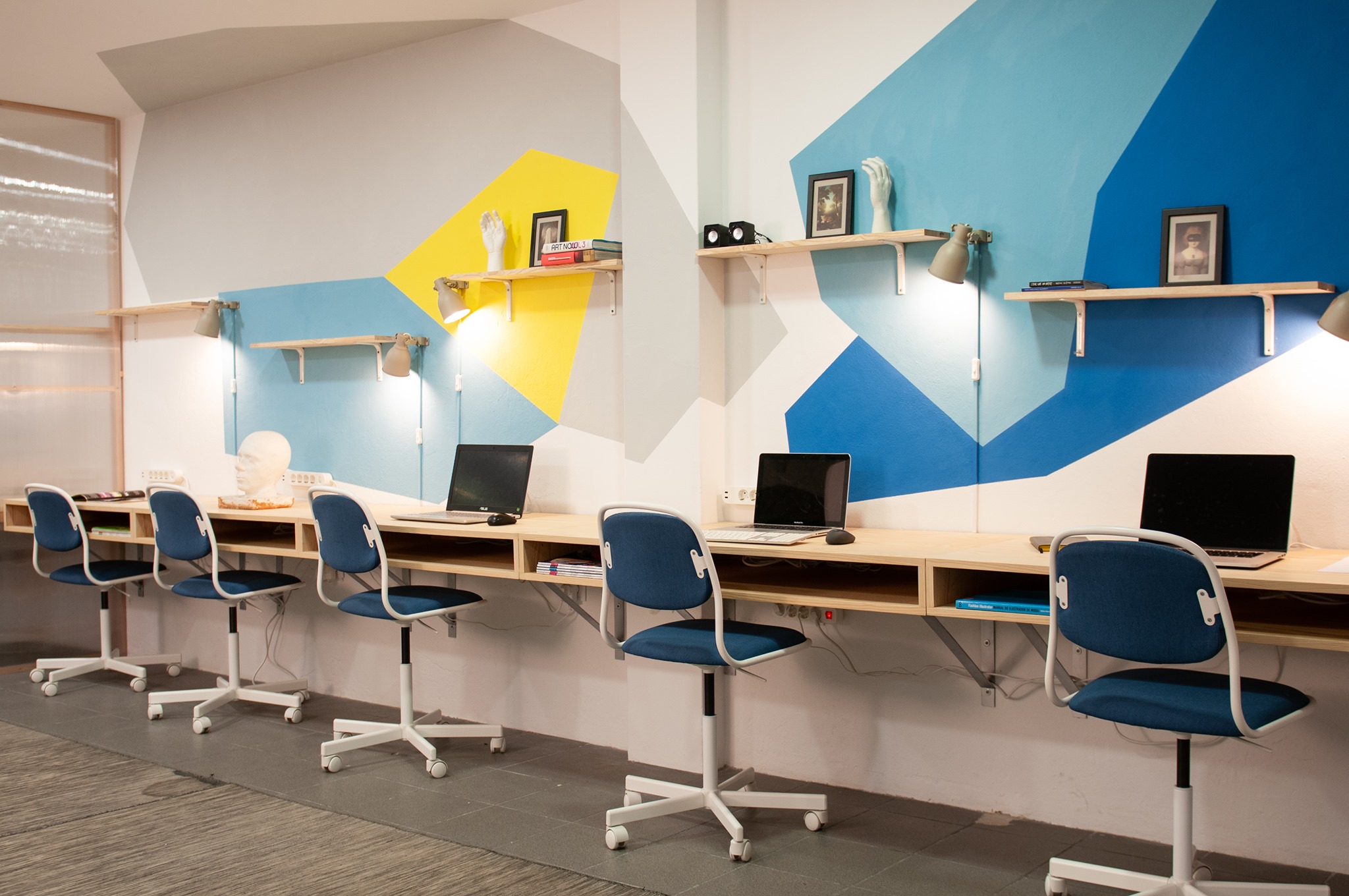 zdjęcie przedstawia salę z rzędem biurek, na których stoją laptopy oraz przypisane do nich krzesła biurowe. Na ścianie widać różnokolorowe wzory, w odcieniach niebieskiego i żółci, a także przytwierdzone do ściany półki.
